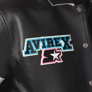Puede incluir: Chaqueta de cuero negra con el parche del logo "AVIREX" y "S". El parche presenta el nombre de la marca en azul y negro, con un contorno blanco y rosa. La chaqueta tiene cierre de bot&oacute;n a presi&oacute;n y una raya blanca en la manga.