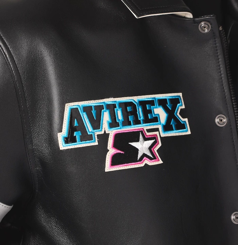 Puede incluir: Chaqueta de cuero negra con el parche del logo "AVIREX" y "S". El parche presenta el nombre de la marca en azul y negro, con un contorno blanco y rosa. La chaqueta tiene cierre de bot&oacute;n a presi&oacute;n y una raya blanca en la manga.