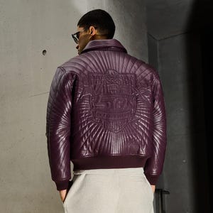 May include: A quilted, deep purple leather jacket with a ribbed collar and cuffs. The back features an embossed design with the text "VIBE 50". The jacket is paired with light gray pants.