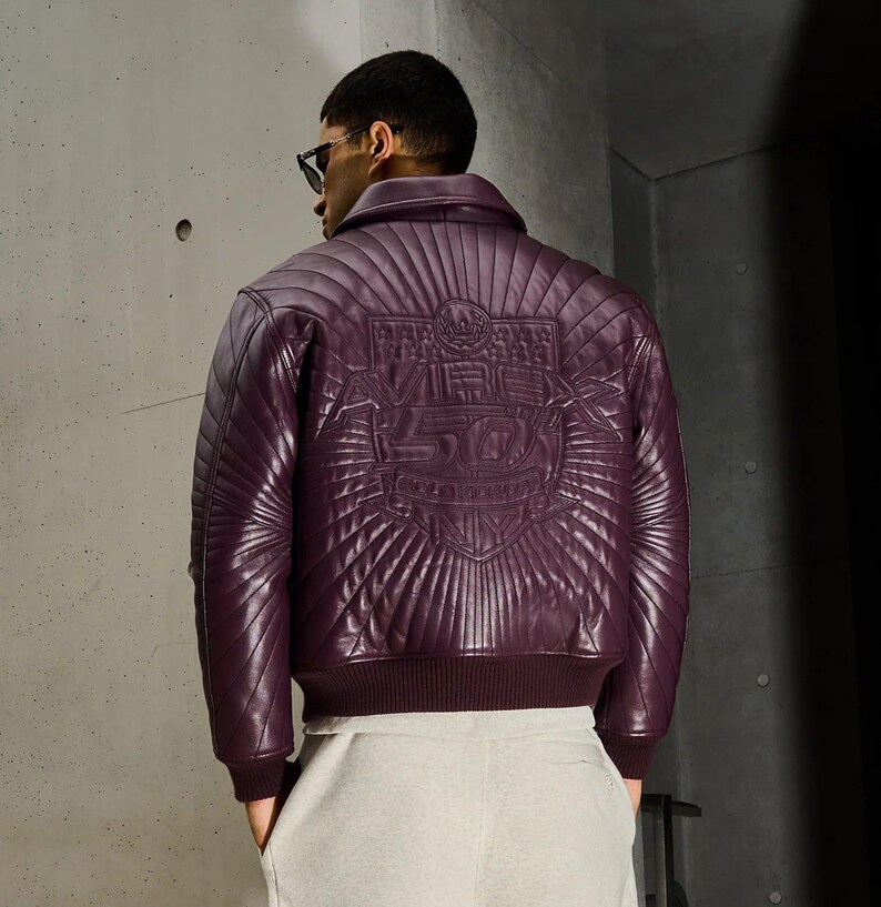 May include: A quilted, deep purple leather jacket with a ribbed collar and cuffs. The back features an embossed design with the text "VIBE 50". The jacket is paired with light gray pants.