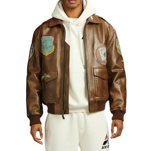 May include: Brown leather bomber jacket with patches, a zip closure, and two front pockets. The jacket has a ribbed collar and cuffs. The patches include military-style emblems and text. The model is wearing a white hoodie underneath.
