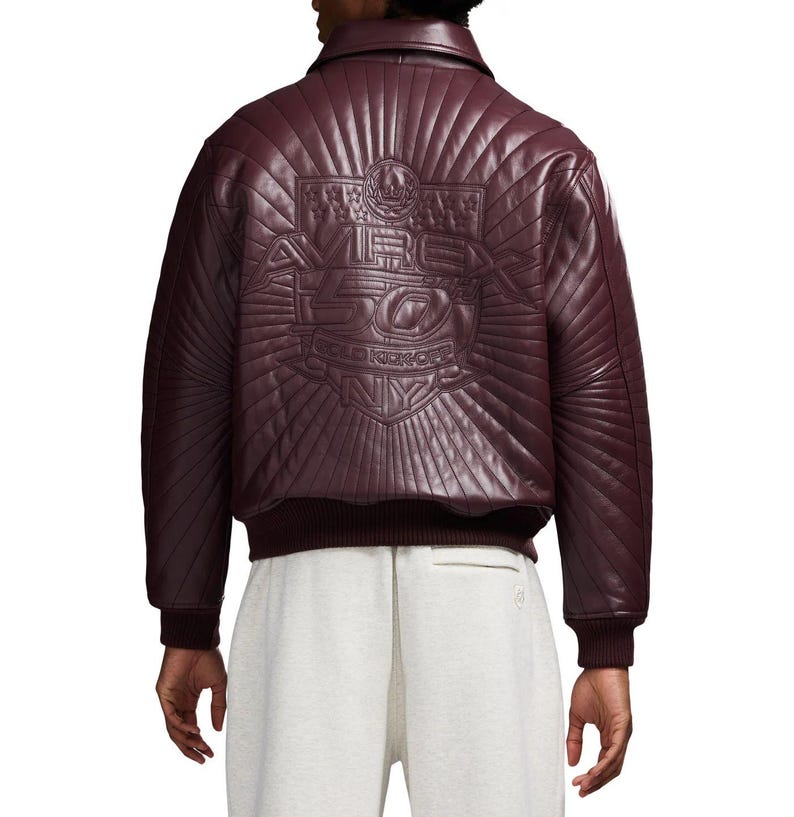 May include: Burgundy leather bomber jacket with quilted design and ribbed cuffs. The back features an embossed graphic with the text "AVIREX 50 GOLD KICKOFF NY". The jacket has a classic collar and a dark burgundy ribbed hem.