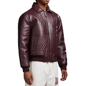 May include: A burgundy leather jacket with a zip-up front, a collar, and a ribbed hem and cuffs. The jacket features a sunburst design and the word "AMIRI" stitched across the chest.
