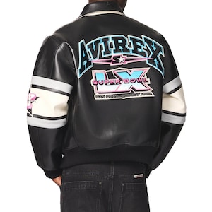 Puede incluir: Chaqueta bomber de cuero negro con mangas a rayas blancas y grises. La parte trasera presenta el texto "AVIREX" en azul y rosa, con "SUPER BOWL LX SAN FRANCISCO BAY AREA" debajo. La chaqueta tiene un dobladillo y cuello acanalados.
