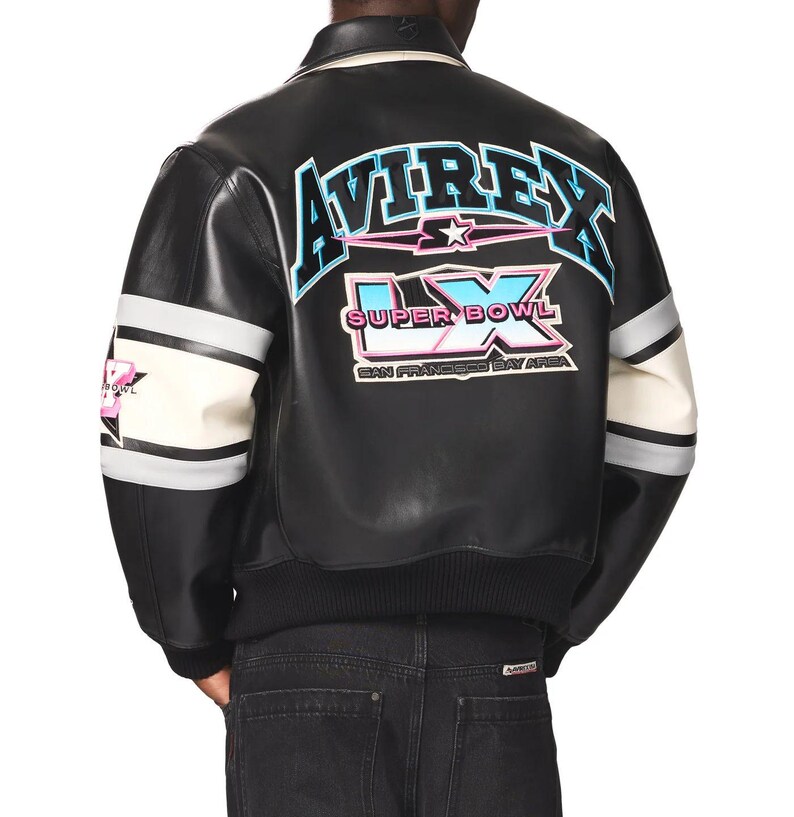 Puede incluir: Chaqueta bomber de cuero negro con mangas a rayas blancas y grises. La parte trasera presenta el texto "AVIREX" en azul y rosa, con "SUPER BOWL LX SAN FRANCISCO BAY AREA" debajo. La chaqueta tiene un dobladillo y cuello acanalados.
