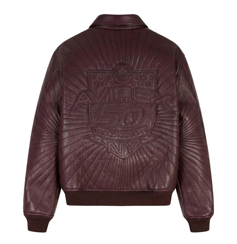 May include: A burgundy leather bomber jacket with a quilted design. The back features an embossed logo with the text "AVIREX 50 GOLD KICKOFF NY" in a sunburst pattern. The jacket has a ribbed hem and cuffs.