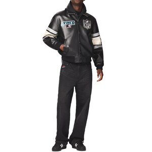 Puede incluir: Chaqueta de cuero negra con rayas blancas en las mangas y el logo de la NFL. La chaqueta tiene cierre de botones y cuello negro. La persona lleva vaqueros negros y zapatillas deportivas negras y blancas.