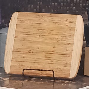 May include: Rectangular bamboo cutting board with rounded corners and a light brown color. The board has a natural wood grain pattern and is supported by a dark metal stand. The edges are a lighter shade of brown.