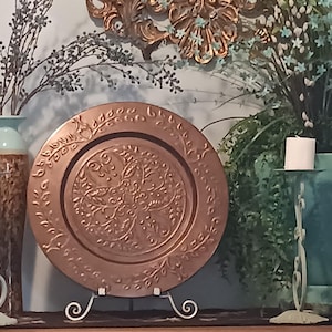 May include: A copper-colored decorative plate with an embossed floral design, displayed on a white metal stand. The plate is surrounded by decorative elements, including a teal vase, candles, and greenery, creating a warm, inviting ambiance.