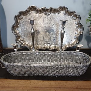 May include: A silver-toned serving tray with ornate detailing, flanked by two tall candlesticks and two smaller candle holders. A woven basket sits in front, with a decorative swan and candelabra visible.