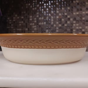 Puede incluir: Un cuenco de cerámica para servir con interior crema y borde marrón. El cuenco presenta un patrón trenzado decorativo marrón alrededor del centro. Ideal para servir alimentos.