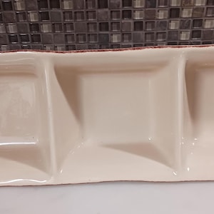 Könnte beinhalten: Cremefarbene rechteckige Servierschale mit drei quadratischen Fächern. Die Schale hat ein Blattdesign an der Seite und einen braunen Rand. Ideal zum Servieren von Snacks oder Vorspeisen. Die Schale ist etwa 30 cm lang.