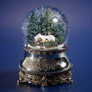 May include: A decorative snow globe featuring a winter scene with a house and evergreen trees. The glass globe sits atop a silver and bronze-toned base with ornate detailing. The scene includes a house with lit windows and a full moon.