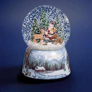 Può includere: Una palla di neve con una scena invernale festosa. Presenta Babbo Natale, un cervo e alberi sempreverdi all'interno di una cupola di vetro trasparente. La base è decorata con un paesaggio invernale con una baita, alberi e montagne.