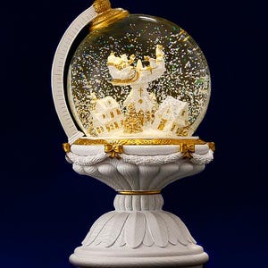 May include: A decorative snow globe featuring a winter village scene with buildings and a church. The globe is clear with floating glitter and sits atop a white and gold pedestal base. The globe is supported by a white arch.