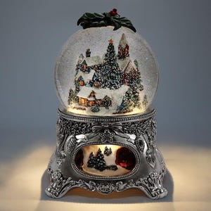 May include: A decorative snow globe with a winter village scene inside. The globe sits on a silver-colored base with ornate detailing. The top of the globe is adorned with green leaves and red berries. The scene inside features snow-covered houses and trees.