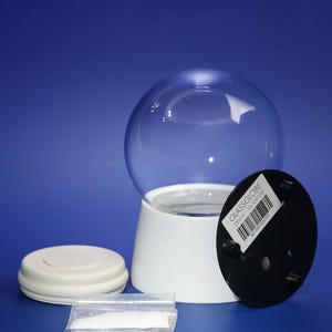 Peut inclure: Un globe de neige démonté avec un dôme en verre transparent, une base blanche et une base noire. Un petit sac en plastique de granulés blancs et un couvercle blanc sont également présents. Le texte "GLASSGLOBE" est sur la base noire.