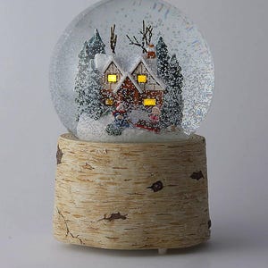 May include: A snow globe featuring a winter scene with a house, trees, and figures on a sled. The house has lit windows. The globe sits on a base that resembles a tree trunk. The snow globe is a festive holiday decoration.