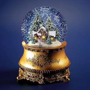May include: A decorative snow globe featuring a winter scene with a cabin, evergreen trees, and falling glitter. The glass globe sits atop a gold-colored, ornate base with a detailed floral design. The background is a deep blue.
