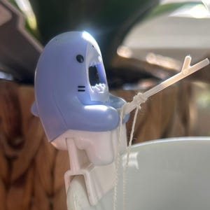 May include: A light blue shark-shaped tea infuser with a white fishing rod. The infuser is designed to hang on the edge of a mug. The shark has black eyes and a wide open mouth. The tea infuser is made of plastic.