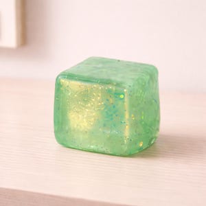 May include: A translucent, square-shaped object with rounded edges. The object is a light green color with gold and teal glitter embedded throughout. It sits on a light-colored wooden surface.