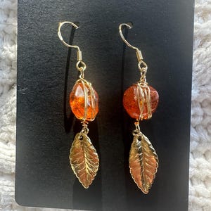 May include: A pair of dangle earrings featuring amber-colored, wire-wrapped beads and gold-colored leaf charms. The earrings hang from gold-colored ear wires and are displayed on a black card.