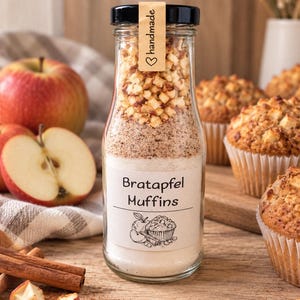 Könnte beinhalten: Eine klare Glasflasche gefüllt mit geschichteten trockenen Backzutaten, mit einem Etikett "Bratapfel Muffins" und einer Illustration von Apfel und Muffin. Ein beigefarbener Anhänger mit der Aufschrift "handmade" ist am schwarzen Deckel befestigt.
