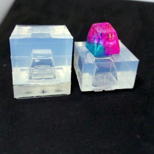 May include: Two clear silicone molds for a custom keyboard keycap. One mold is empty, the other holds a colorful keycap with pink, blue, and purple hues. The keycap has a glossy finish and is rectangular.