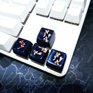 May include: Close-up of custom keyboard keycaps with a koi fish design. The keycaps are dark blue with a glossy finish, and each features a unique arrangement of red and white koi fish. The keycaps are on a white keyboard.