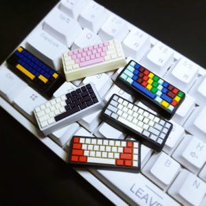 May include: A collection of miniature keyboard keycap designs in various color schemes, including pink, blue, red, and black. The keycaps are displayed on a white keyboard, showcasing their different layouts and aesthetics. The keycaps are a unique accessory for mechanical keyboards.