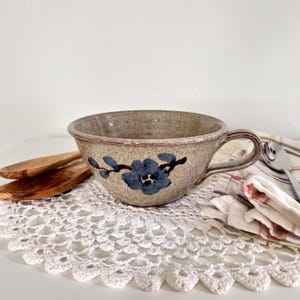 May include: A speckled beige ceramic soup bowl with a single handle, featuring a hand-painted blue flower design on the front. The bowl has a wide, flared rim and a smooth, glazed finish, showcasing a natural, earthy aesthetic with subtle brown flecks.