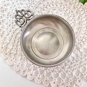 May include: A small, antique pewter bowl with an ornate handle. The bowl has a circular base and a decorative handle with a scroll design. The bowl is sitting on a white, crocheted doily.