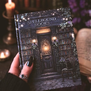Puede incluir: Un libro de tapa dura titulado "Spellbound Books" con una ilustración detallada de una entrada a una biblioteca mística. La portada representa una puerta oscura, estanterías y un gato negro en una silla, con iluminación suave y detalles florales. El libro se sostiene en una mano.
