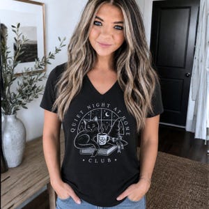 Puede incluir: Una camiseta negra con cuello en V que presenta un estampado gráfico blanco de un gato sentado sobre un libro junto a un sombrero de bruja y una taza humeante con un pentagrama. El texto dice Quiet Night At Home Club bajo un arco de ventana.
