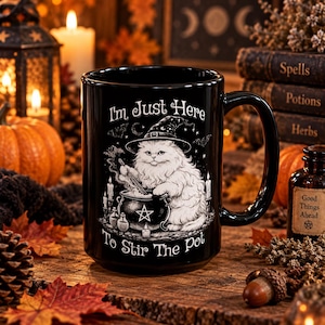 May include: A black ceramic mug featuring a white illustration of a fluffy cat wearing a witch hat and stirring a cauldron. The text "I'm Just Here To Stir The Pot" is printed in white above and below the cat, surrounded by small stars and bats.