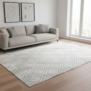 May include: A modern living room featuring a light gray sofa with matching pillows, a geometric patterned rug, and a black floor lamp. The rug has a repeating square design in shades of gray and white. Two framed abstract art pieces hang on the wall.