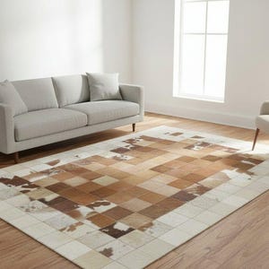 Può includere: Un tappeto rettangolare con un design patchwork nei toni del marrone, beige e crema. Il tappeto è posato su un pavimento in legno in un soggiorno, con un divano grigio e una poltrona beige.