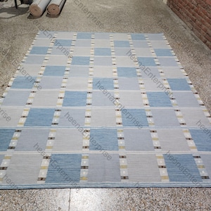 Puede incluir: Una alfombra rectangular con un patrón geométrico. La alfombra presenta cuadrados alternos de color azul claro y gris, con finas rayas verticales amarillas y marrones. La alfombra está hecha de un material tejido.