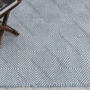 Puede incluir: Alfombra con estampado geométrico azul y blanco con diseño de rombos repetidos. Una pequeña mesa auxiliar de madera con un libro encima se encuentra en la esquina superior izquierda.