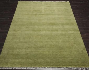 May include: A rectangular, solid green rug with a fringe on the bottom edge. The rug is made of a soft material and is placed on a dark brown wooden floor. The rug's color is a muted, earthy green.