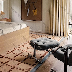 Op de afbeelding: Slaapkamerinterieur met een laag houten bed, een patroonkleed met bruine geometrische vormen en een grijze gestoffeerde poef met een chromen frame. Een abstract schilderij hangt aan de muur.