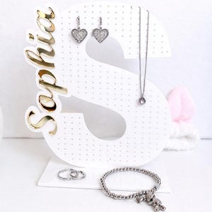 May include: A white jewelry display stand in the shape of the letter "S" with the name "Sophia" in gold script. The stand holds heart-shaped earrings, a necklace, rings, and a beaded bracelet with a teddy bear charm.