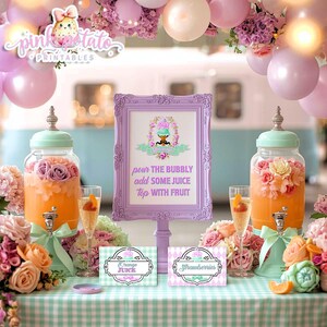 May include: A party scene with a light blue and white backdrop, featuring a table with drinks and decorations. Two glass beverage dispensers with floral arrangements flank a framed sign that reads "pour the bubbly add some juice top with fruit."