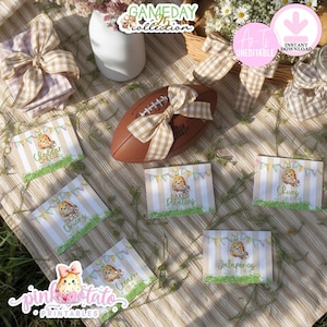 May include: A picnic setting featuring a brown American football, food labels, and decorative items. The labels display text such as "Cheddar Cheese," "Diced Onions," "Cream," "Baked Potatoes," "Chives," and "Jalapeños." The image includes text that says "Gameday Collection" and "Pink Potato Printables."