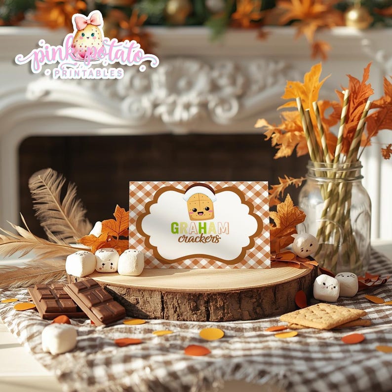 May include: A rustic wooden slice displays a "Graham Crackers" card with a cartoon cracker illustration. The scene includes chocolate bars, marshmallows, and fall foliage, creating a cozy autumn theme. A jar with fall leaves and decorative elements complete the arrangement.