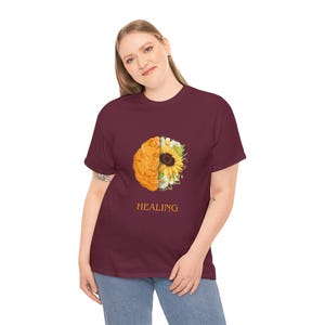 Può includere: T-shirt bordeaux con un disegno grafico di un cervello e un girasole, con la scritta "HEALING" stampata sotto. Il cervello è arancione e giallo, mentre il girasole è giallo e bianco. La t-shirt è indossata da una persona.