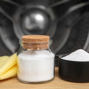 Puede incluir: Un frasco de vidrio transparente con tapa de corcho lleno de polvo blanco, junto a una taza medidora negra también llena de polvo blanco. Un paño amarillo está a la izquierda. El fondo es un tambor oscuro de lavadora.