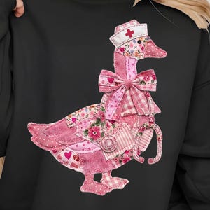May include: Black sweatshirt featuring a patchwork goose design. The goose wears a nurse's cap with a red cross and a pink bow. The goose has a stethoscope around its neck, and the design incorporates floral and heart patterns.