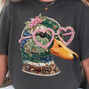 May include: Dark gray t-shirt featuring a whimsical duck design. The duck is adorned with heart-shaped glasses, a floral crown, and a patchwork pattern. The design includes a pearl border. The shirt is a relaxed fit.