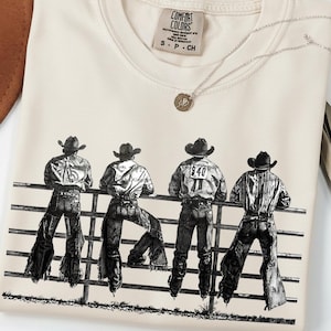 May include: A cream-coloured t-shirt with a black and white illustration of four cowboys leaning on a fence. The shirt has a crew neck and short sleeves. A silver necklace with a small pendant is on top.